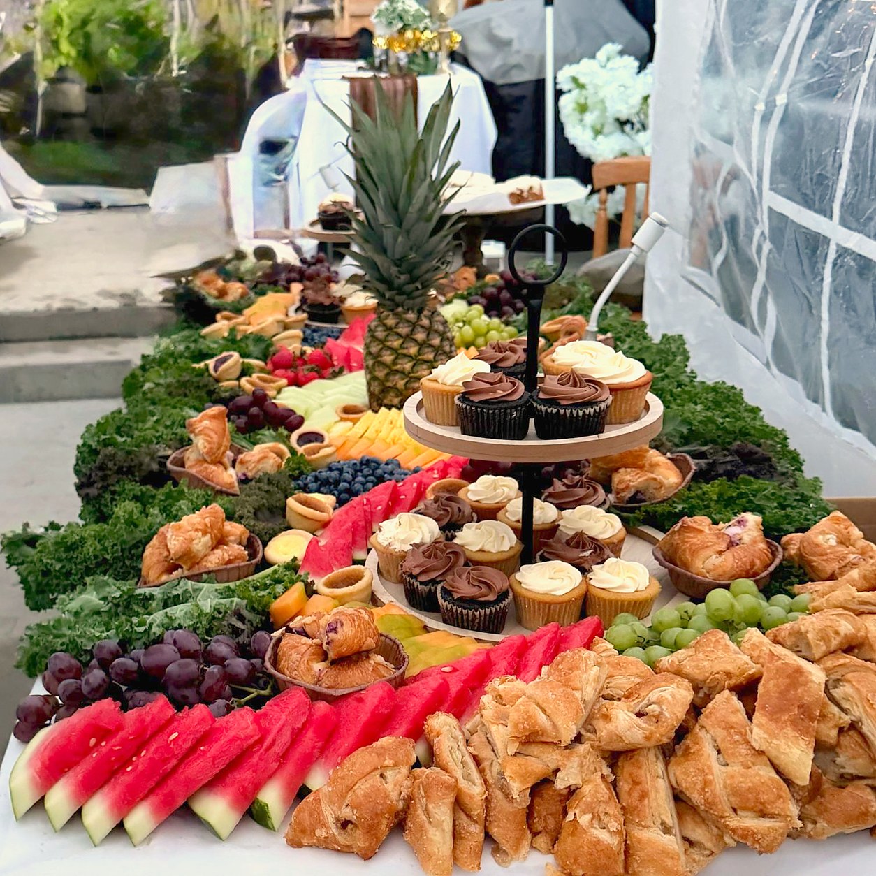 A long grazing table of fruit, pastries, cupcakes and a pineapple centrepiece in a garden tent